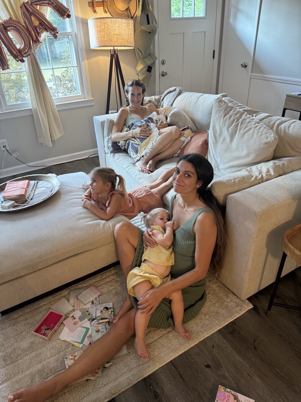 Two mothers nursing their toddlers in a living room while older children play nearby.
