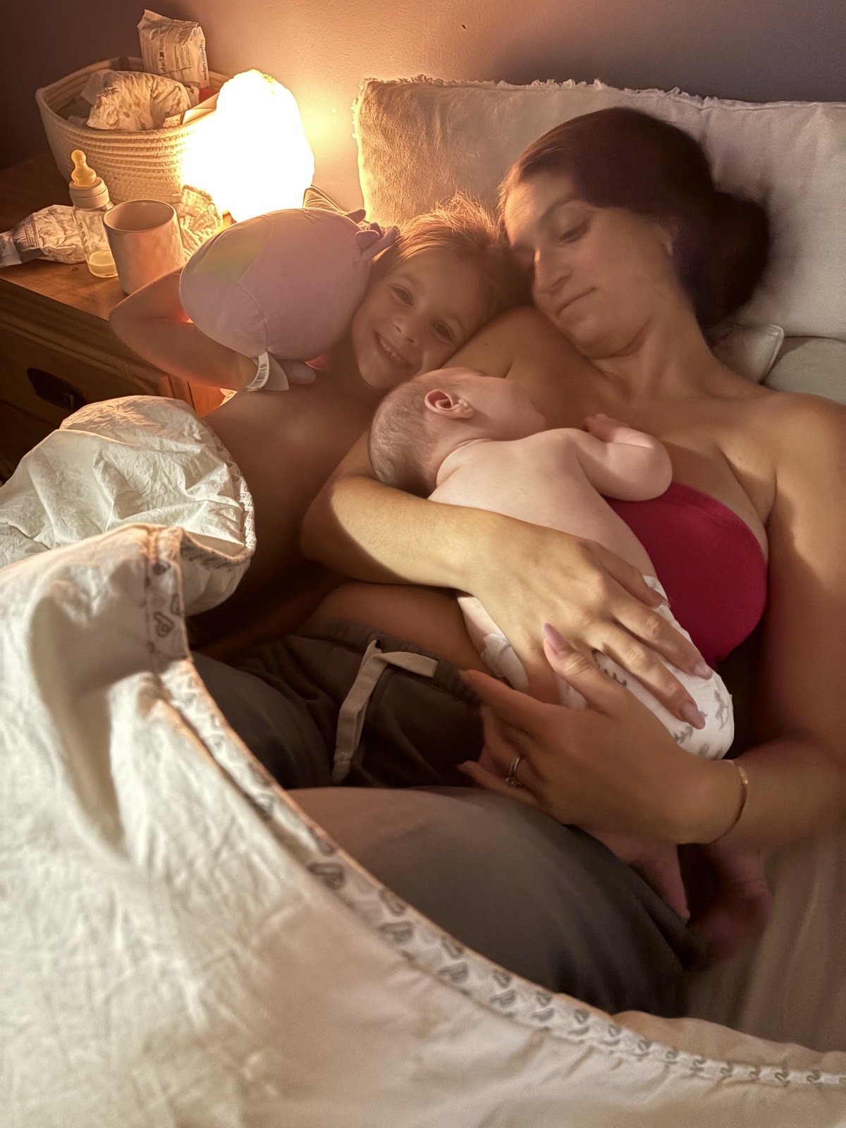 A mother nursing her newborn in bed at night by lamplight while her older child snuggles beside her.